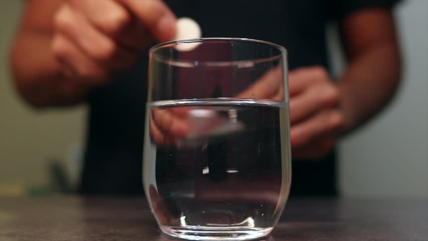 Effervescent white tablet fizzing and dissolving in a glass of water, creating bubbles. The pill is dropped into the water to provide relief from a headache or hangover