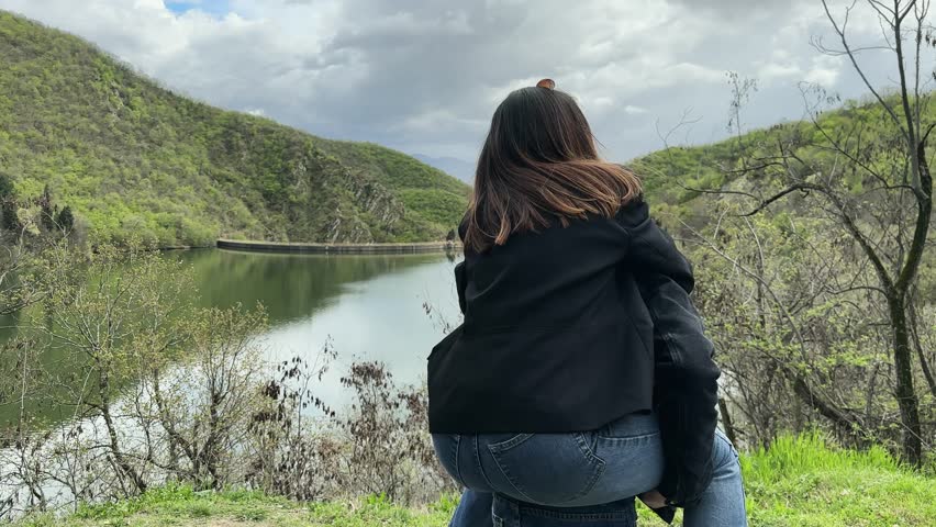 Happy family brother and sister having fun playing piggyback with woman is sitting on man back. She is wearing a black jacket and jeans. The scene is set in a forest with a lake in the background