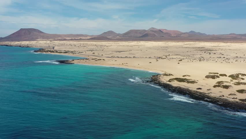 Stunning aerial drone footage of Corralejo Natural Park in Fuerteventura, Canary Islands, showcasing the vast white sand dunes, crystal-clear turquoise Atlantic beaches, and the road crossing the natural landscape towards the iconic Red Mountain volcanic cone.