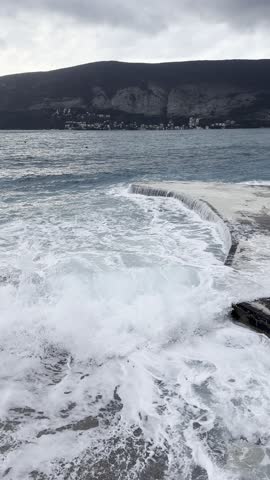 Rough sea waves crashing against the pier and shoreline, creating dramatic splashes and white foam during stormy weather. Powerful water movement and unstable sea conditions along the coast. 