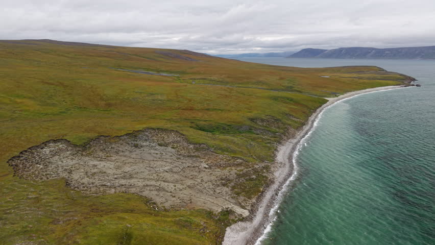 Slow drone flyover reveals a thaw slump on the coastal tundra and emerald waves of Lavrentiya Bay on an overcast summer day. Remote Arctic coastline, pristine beach, travel and documentary friendly