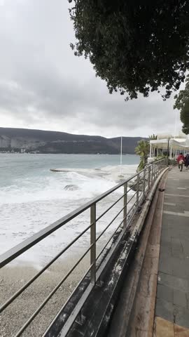 Rough sea waves crashing against the pier and shoreline, creating dramatic splashes and white foam during stormy weather. Powerful water movement and unstable sea conditions along the coast. 