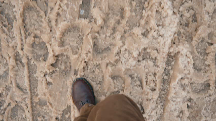 Top-down view of young man wearing brown pants and leather boots walking through slushy snow on city street. Melting snow is mixed with dirt and water, with many footprints visible in snowy slush