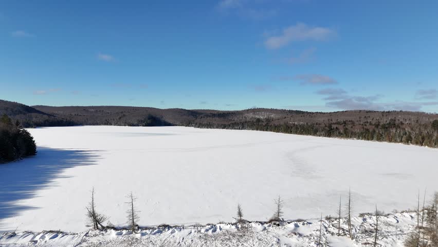 An aerial glides over a deep winter landscape, featuring a frozen Billy Lake at Algonquin Park surrounded by snow-covered evergreen forests under a clear blue sky