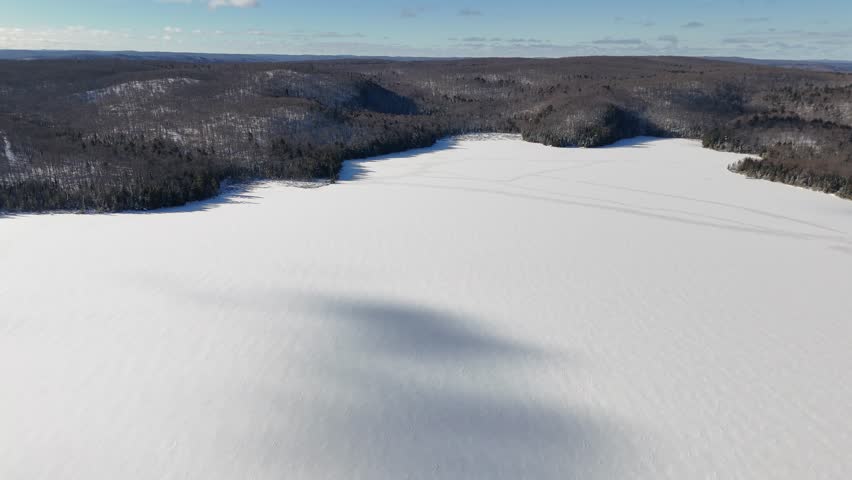 An aerial pulls back to reveal the vast, snow-covered expanse of Billy Lake and the surrounding winter evergreen forests in Algonquin Park, Canada