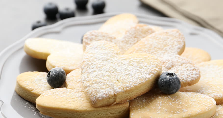 Woman taking tasty heart shaped butter cookie with sugar powder from plate at grey table, closeup