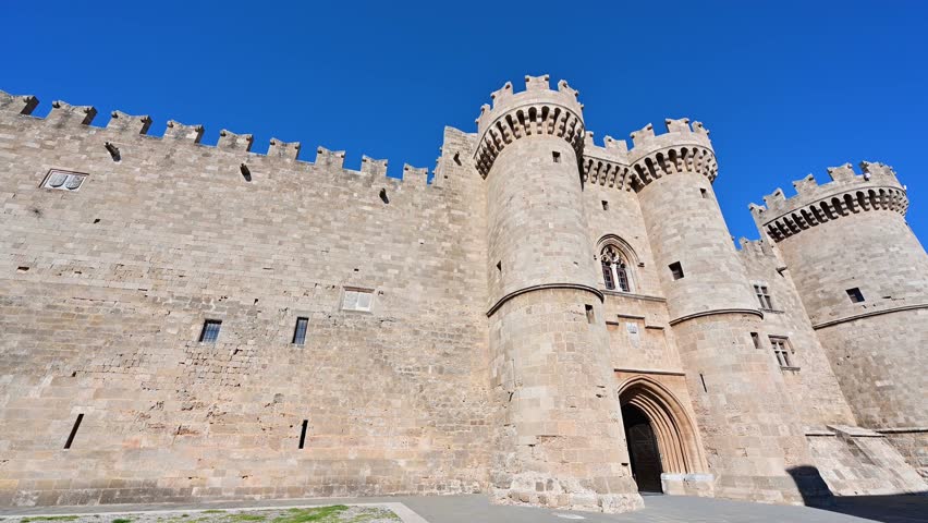 Rightward panning view along the exterior walls of the Palace of the Grand Master of the Knights of Rhodes in Rhodes Old Town, Greece, emphasizing massive stone fortifications and medieval architecture under daylight.