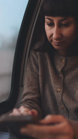 vertical shot. A cheerful young woman texting on a mobile phone on a train. Side view of a woman on a train. A girl using social media. A girl on a train.