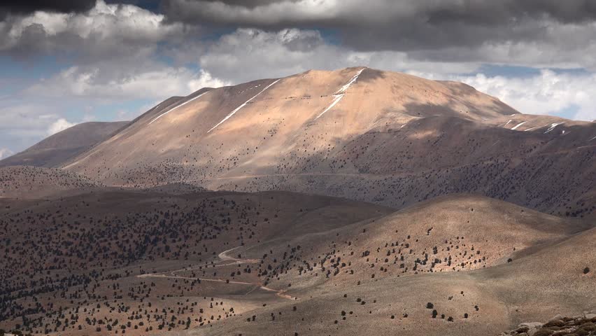Time lapse cloudscape over arid Atlas Mountains with sparse trees under Mediterranean climate. Dramatic light moves across rugged ridges in North Africa showing semi arid mountain landscape.