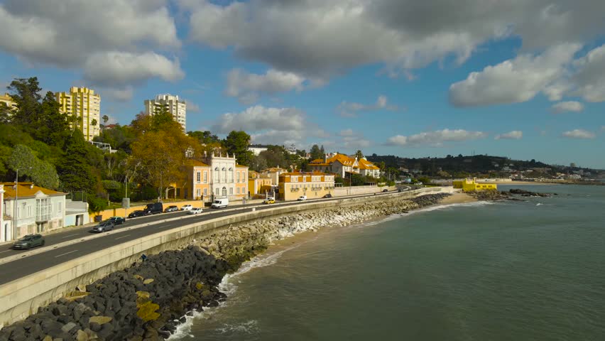Wide aerial orbit along Caxias and Paço de Arcos shoreline with Tagus River, revealing traditional Portuguese coastal homes villas, greenery, moving traffic and ocean under dramatic clouds near Lisbon