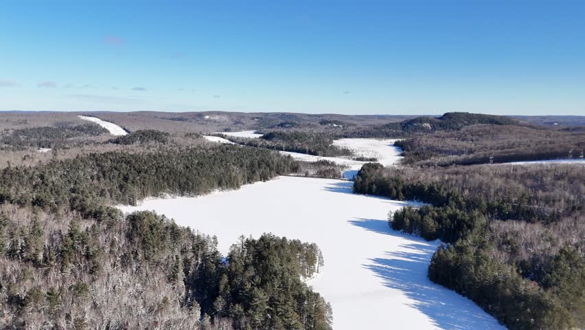 Aerial approaches a vast, snow-covered Oram Lake surrounded by dense evergreen forests and rolling hills in Algonquin Provincial Park on a sunny winter day