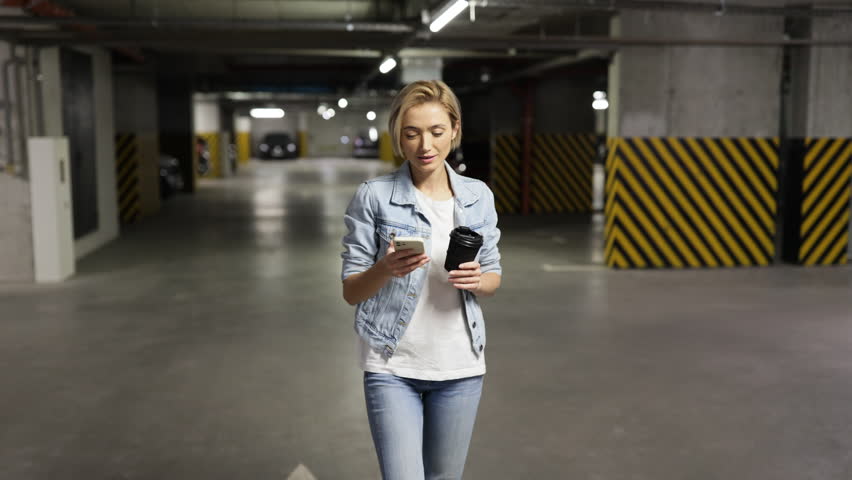 A woman walks through a parking garage while talking on her phone and holding a coffee cup