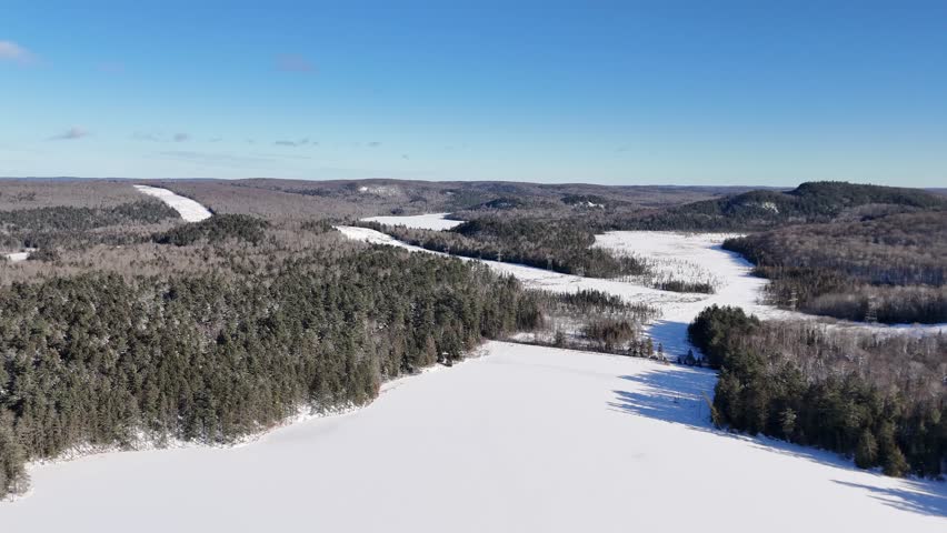 An aerial drone shot approaches a vast, snow-covered Oram Lake surrounded by dense evergreen forests and rolling hills in Algonquin Provincial Park on a sunny winter day