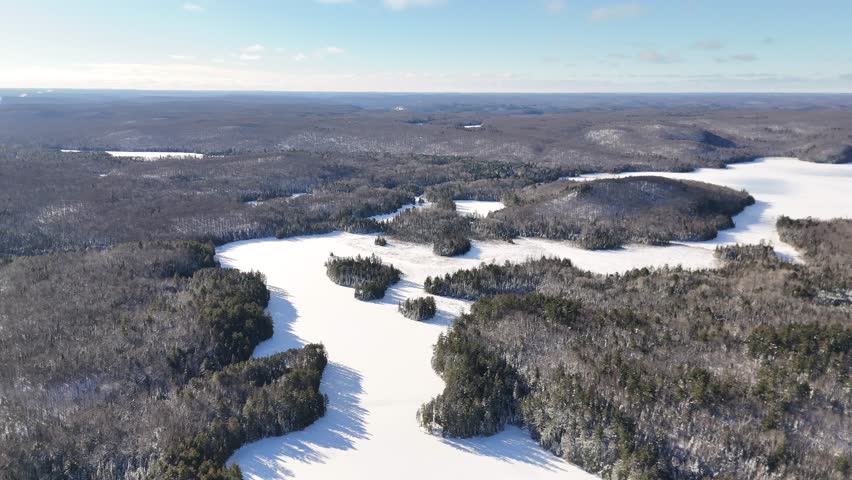 An aerial approaches a vast, snow-covered Oram Lake surrounded by dense evergreen forests and rolling hills in Algonquin Provincial Park on a sunny winter day