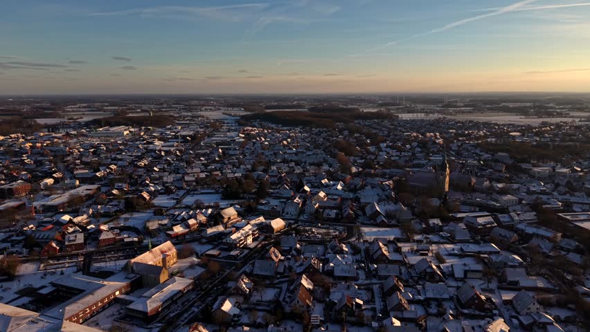Aerial establishing shot of snowy small town in American during golden hour. Historic church tower in snowy village. Wide shot. Sunset time Farmland in distance. Peaceful and quiet residential area.