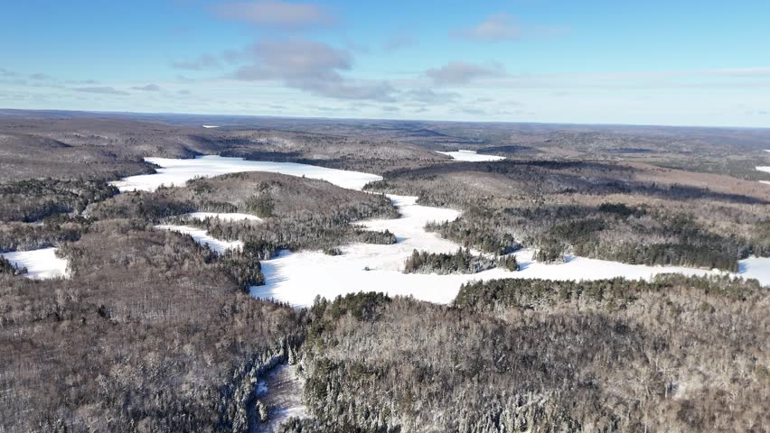 An aerial rises vertically to reveal a panoramic, snow-covered Billy Lake surrounded by dense evergreen forests and rolling hills in Algonquin Provincial Park on a sunny winter day