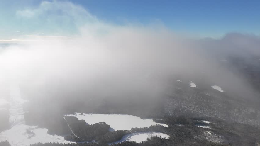 Aerial flight through clouds over snow-covered forests and frozen lakes in Algonquin Provincial Park, Ontario, Canada on a sunny day. Serene winter wilderness landscape