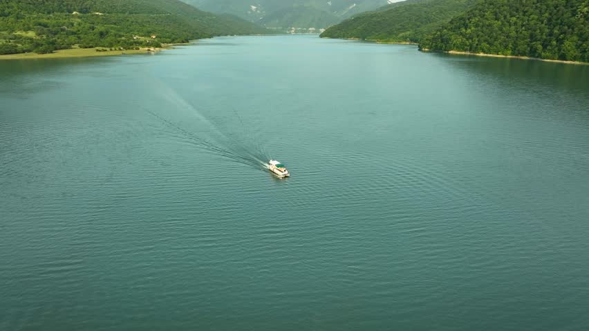 Aerial view of a small boat sailing across a tranquil mountain lake in Georgia, surrounded by green hills and untouched nature.