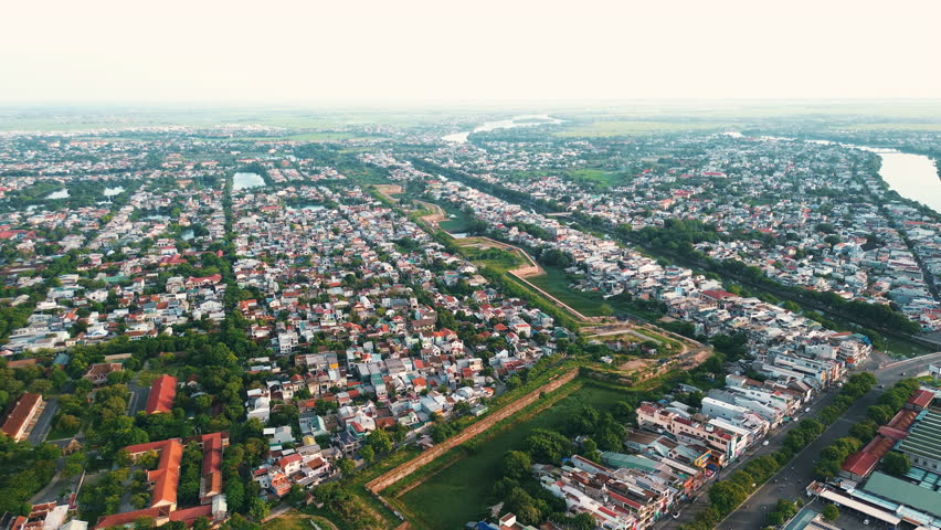 A wide panoramic drone shot showcasing the historic stone walls of the Hue Imperial City (Citadel) integrated within the dense residential landscape and greenery of the modern city.