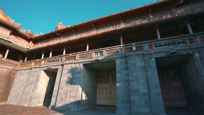 A panoramic low-angle shot of the Noon Gate at the Hue Citadel, showing the grand U-shaped stone foundation and the ornate wooden pavilion on top under a clear blue sky.