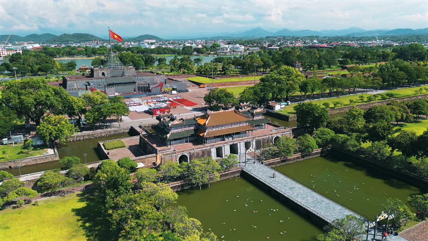 A stunning high-angle drone shot capturing the grand entrance of the Hue Citadel, featuring the Noon Gate (Ngo Mon), the massive Ky Dai (Flag Tower) with the Vietnamese flag, the surrounding moat, and