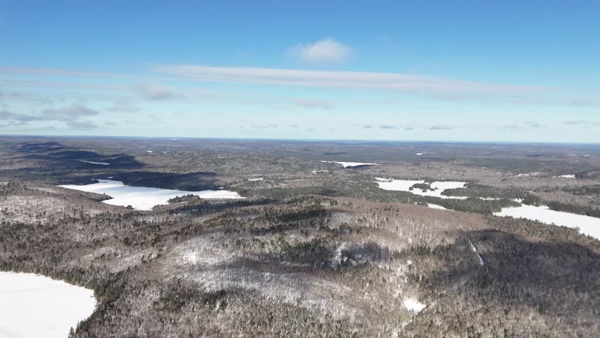 An aerial captures a wide landscape as it descends towards a snow-covered Oram Lake surrounded by dense evergreen forests and rolling hills in Algonquin Provincial Park