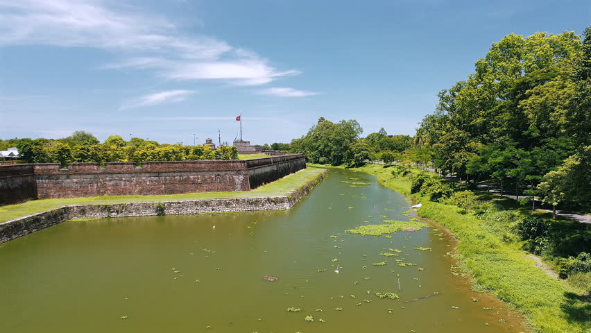 A steady drone shot showcasing the ancient stone walls and the surrounding green moat of the Hue Citadel, with the Ky Dai (Flag Tower) and the Vietnamese flag visible under a blue sky.