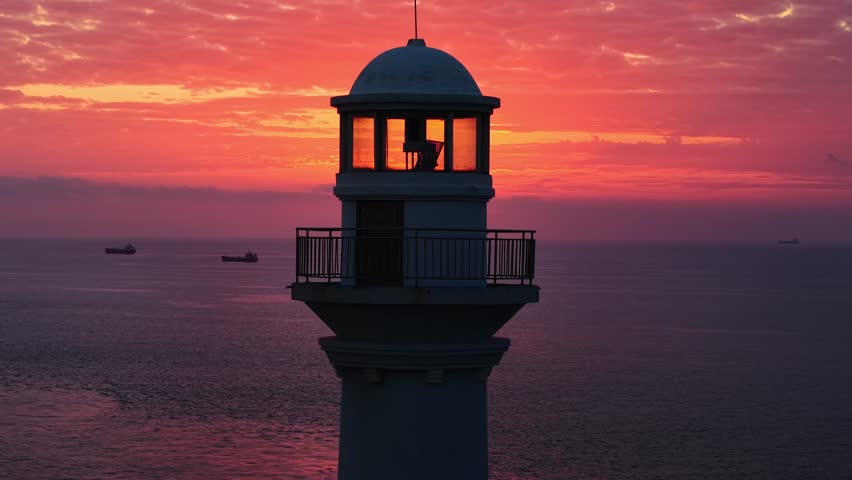 Captivating lighthouse silhouette against ocean sunset with passing cargo ships.