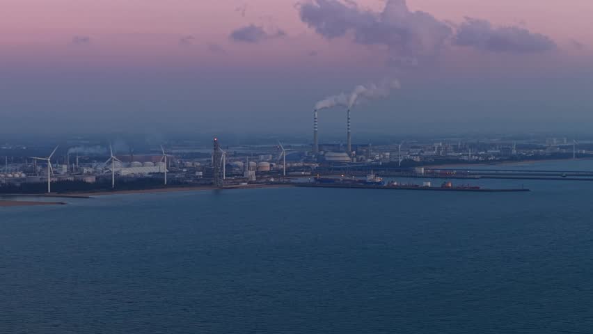 Twilight industrial chemical oil plant and wind turbines and smoke plumes over tranquil sea.