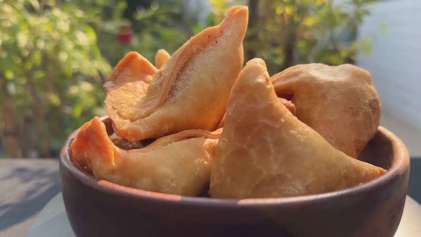 Rotating turntable closeup shot of golden crispy samosas stacked in a bowl, flaky pastry edges catching warm sunlight as gentle motion reveals texture and shape