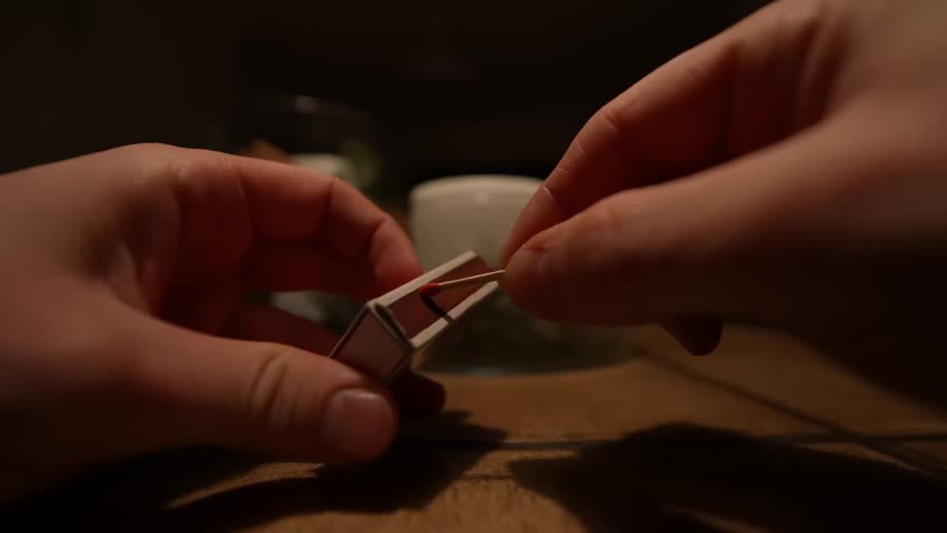Slow motion close-up of hands striking a match, bright flame and drifting smoke in warm light—concept for emergency, indoor, safety, and ignition.