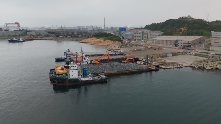 An aerial view captures a bustling industrial port, showcasing heavy machinery operations. In the foreground, multiple barges and tugboats are docked, with excavators actively loading large piles of grey rocks onto a barge. The background features extensive industrial buildings, towering cranes, and smokestacks emitting steam, all set against a backdrop of green hills and an overcast sky. Concept of industrial development and maritime logistics.