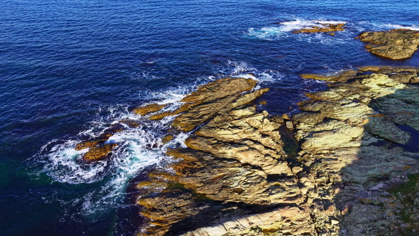 Rough rock formations jut out from the deep blue sea as gentle waves lap against their edges. The sun low on the horizon casts a warm glow over the scene, enhancing the beauty of nature