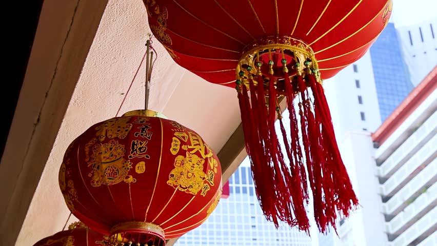Vibrant red lanterns with gold tassels sway gently under a walkway with city buildings behind.