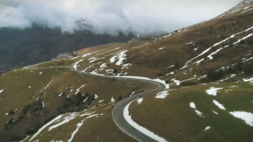 Cinematic aerial tilt-down revealing a winding mountain road near the French border in Larra-Belagua. Snow patches and vegetation dot the landscape under heavy winter clouds.