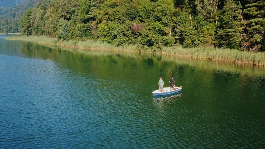 Aerial drone footage of two people fishing from a small boat on a calm forest lake, peaceful outdoor angling scene, friendship, patience, slow living, summer nature retreat, freshwater fishing lifestyle, tranquility and mindfulness in wilderness