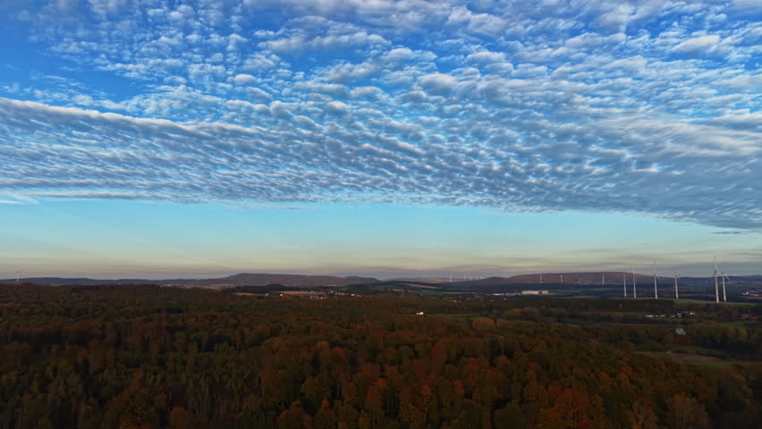The sun sets over a picturesque countryside painting the sky with soft clouds while colorful autumn foliage covers the ground Wind turbines stand tall in the distance.