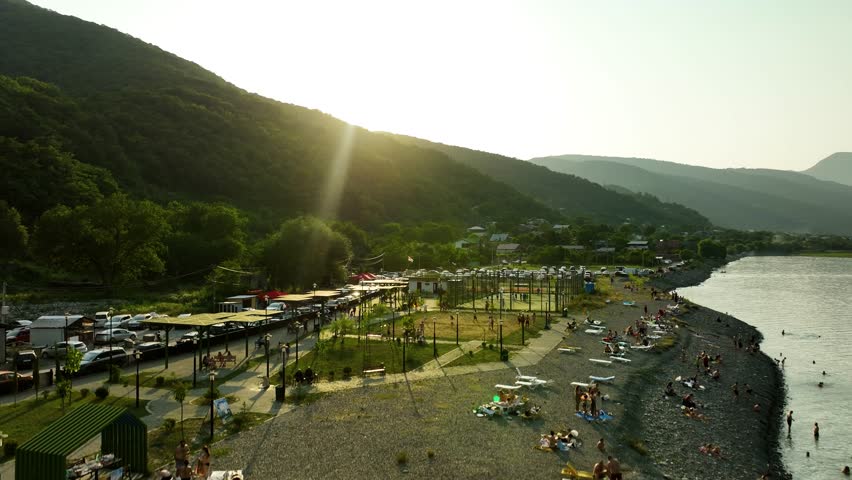 Aerial view of a lakeside village at sunset in Georgia, surrounded by forested mountains and calm water, captured in warm evening light.