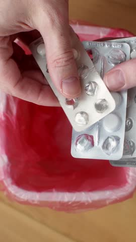 Hands toss leftover tablets in blister packs into red trash bin during home first aid kit cleanup routine and safety check.