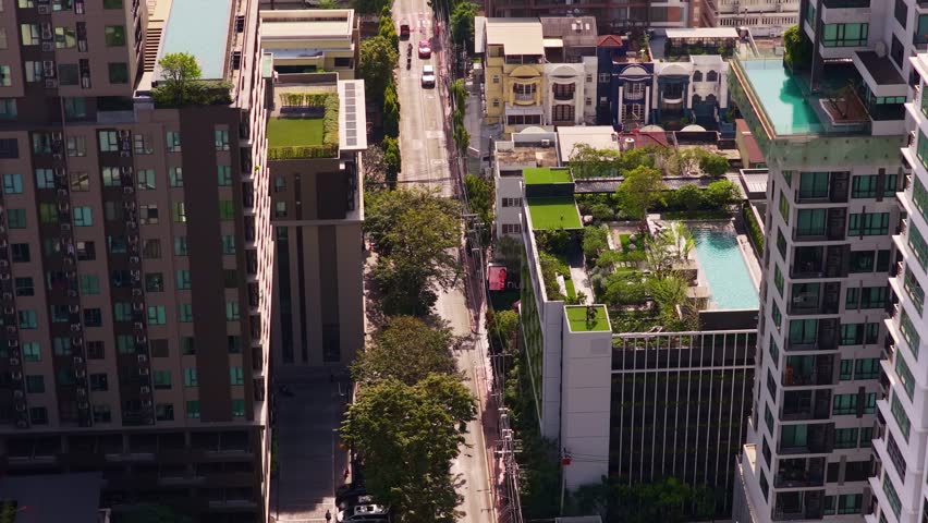 Aerial view looking down on a quiet residential street in Bangkok, Thailand, with cars driving between modern buildings with rooftop pools
