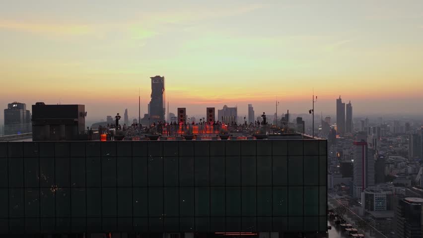Majestic aerial view of the Bangkok skyline at dusk. Young people dance and drink on terrace of modern skyscraper rooftop bar