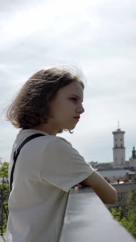 Vertical video. Slow motion, wind blows the hair of a teenage girl who stands high on a terrace or balcony and looks into the distance at the city.