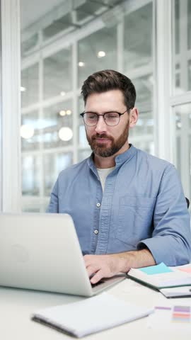 Smiling businessman working on a laptop sitting at workplace in business office. Handsome bearded male worker typing on computer, texting a client, chatting online or busy with project. Vertical video
