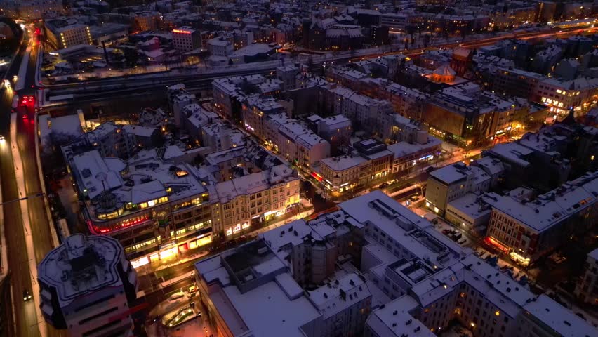 urban shopping street architecture and illuminated streets of Berlin Steglitz district covered in fresh snow during snowy winter evening. Spectacular aerial view tilt up panorama overview drone