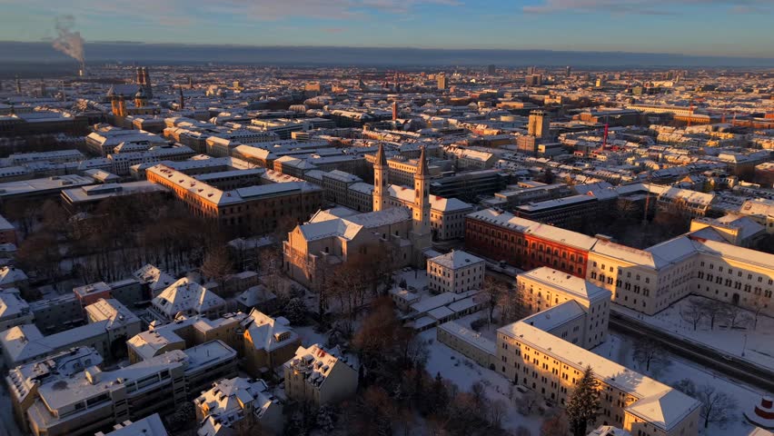 Aerial winter sunrise over Ludwigstrasse in Maxvorstadt, Munich, Germany. Snow-covered rooftops and the twin towers of St. Ludwig Church near LMU glow in soft morning light, cinematic cityscape