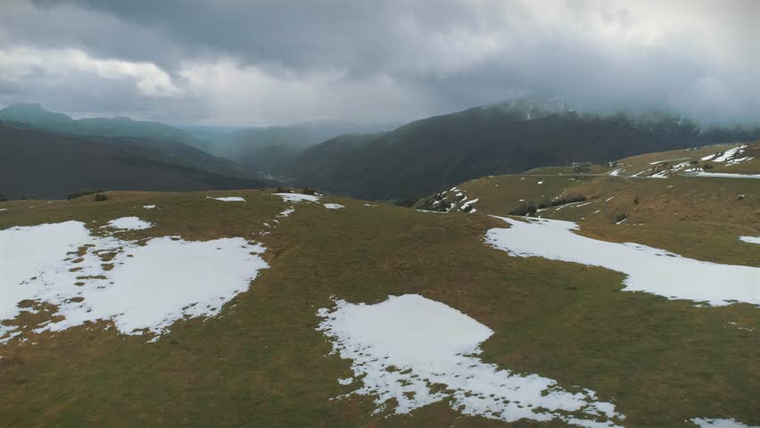 Downward tilting aerial shot over snow-patched mountains in Larra-Belagua, Spain. The drone approaches a cliff to reveal a deep valley with trees, vegetation, and fog.
