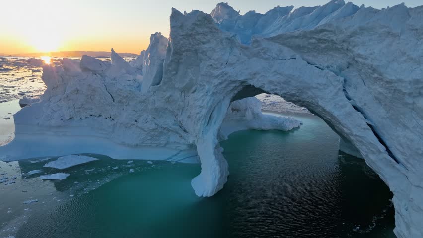 Drone flying forward and rising over a natural ice arch in Disko Bay near Ilulissat, Greenland, frozen Arctic sea with low sun reflection