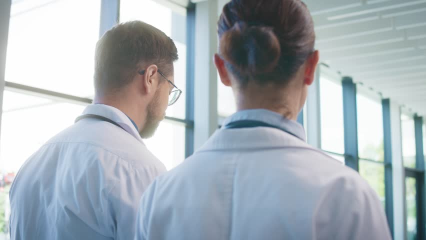 Close camera view from behind two focused doctors walking through bright clinic hallway. Professionals talking casually while moving toward duties. Team preparing for busy day together.