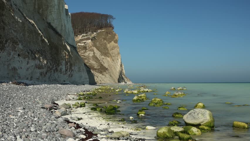 Panoramic view from the white chalk cliffs of Møns Klint overlooking a dramatic rock formation and turquoise sea below. Scenic coastal landscape of Denmark with crystal-clear water and iconic limestone cliffs rising above the shoreline. Natural daylight panorama showcasing the beauty of the Danish coastline and Scandinavian nature. Ideal for travel, tourism, nature, geology, coastal landscapes, and tranquil outdoor concepts.