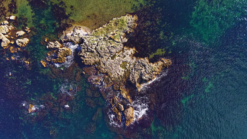 Bright sunlight illuminates the rocky coastline, showcasing a beautiful interplay of land and sea. Crystal-clear waters ripple around the ancient formations, hinting at hidden treasures beneath.